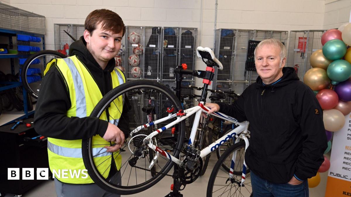 Two men standing in a bicycle repair shop. A white road bike is being held on a repair stand.