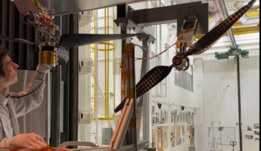 A man in a white clean room suit inspects a horizontal three-bladed rotor. To the right, a vertical two-bladed rotor with a checkered pattern is mounted. Both sit within a large, white industrial testing chamber filled with scaffolding and equipment.
