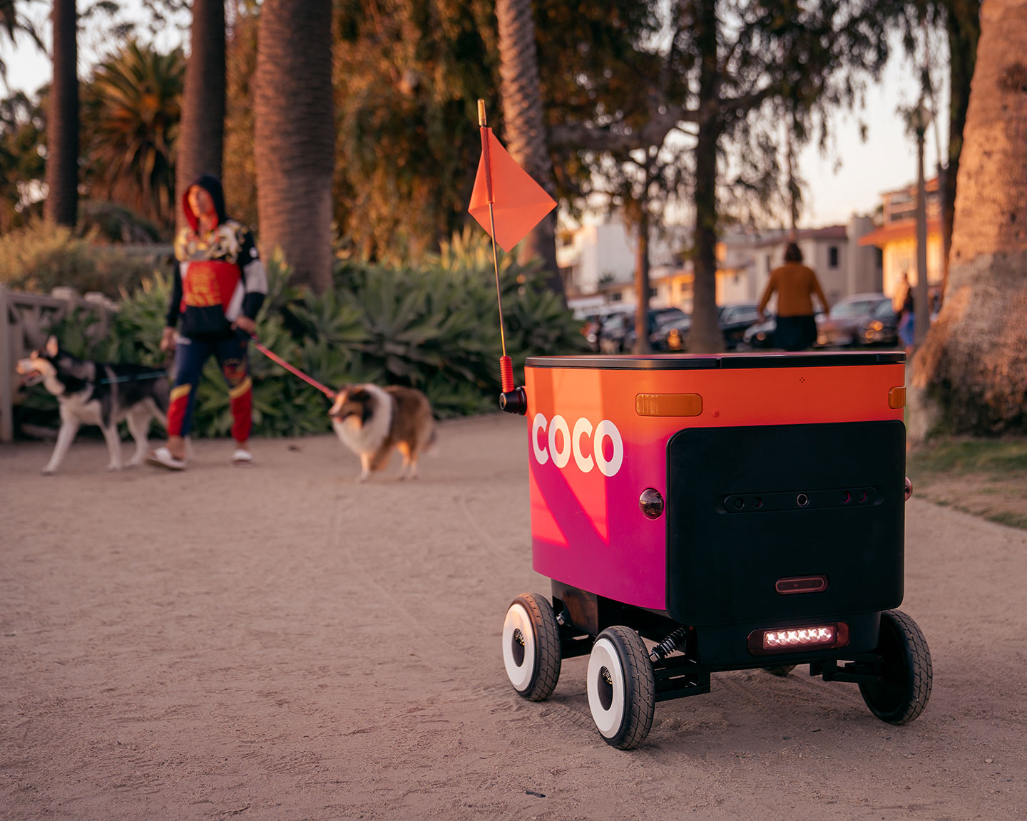 A bright orange and pink Coco delivery robot with a small red flag navigates a sandy park path at sunset, while a person walks two dogs in the background among palm trees.