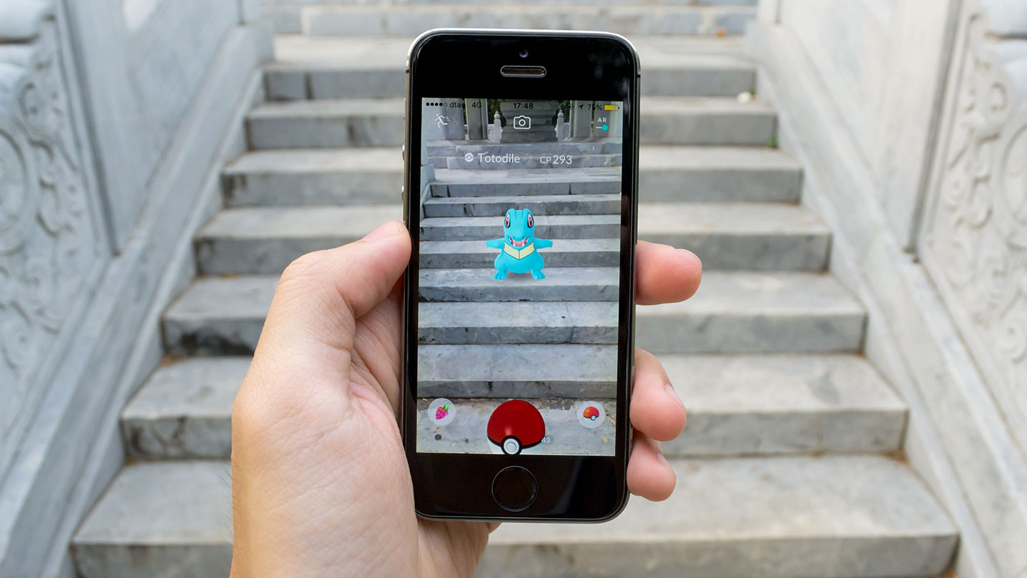 A hand holds a smartphone displaying the Pokémon Go app in augmented reality mode, showing the water-type Pokémon Totodile appearing on a set of stone steps.