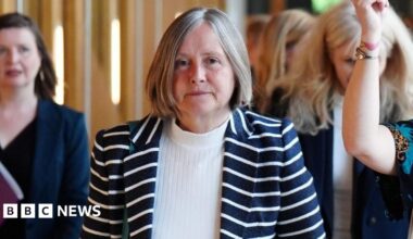 Helen McDade, who has short grey hair, looks at the camera as she walks along a corridor. She is wearing a stripy top over a white jumper