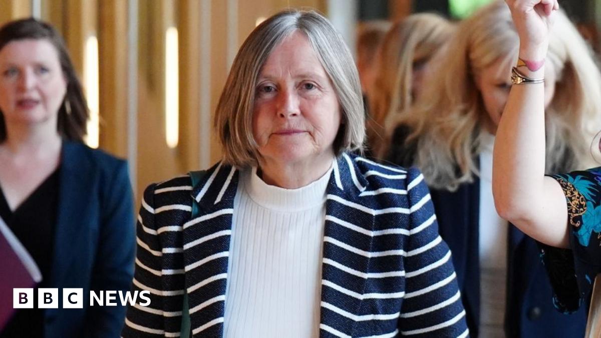Helen McDade, who has short grey hair, looks at the camera as she walks along a corridor. She is wearing a stripy top over a white jumper