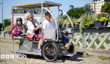 The image shows three people on a Vhélio, with flowers alongside the path in the background.