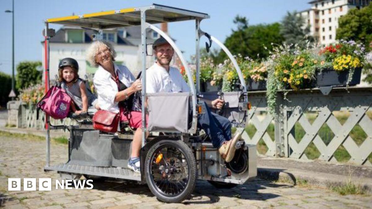 The image shows three people on a Vhélio, with flowers alongside the path in the background.