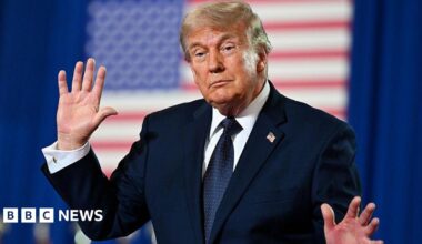 Trump holds up his hands while speaking at an event. He is wearing a dark suit and standing in front of an American flag
