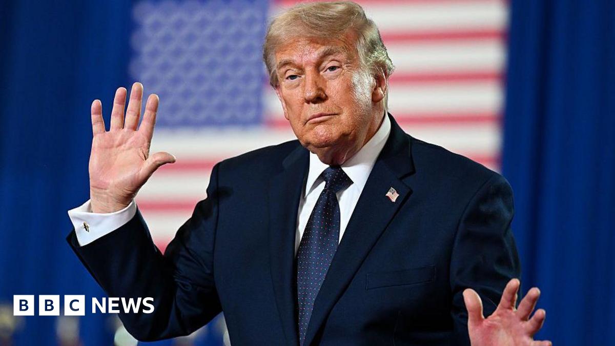 Trump holds up his hands while speaking at an event. He is wearing a dark suit and standing in front of an American flag
