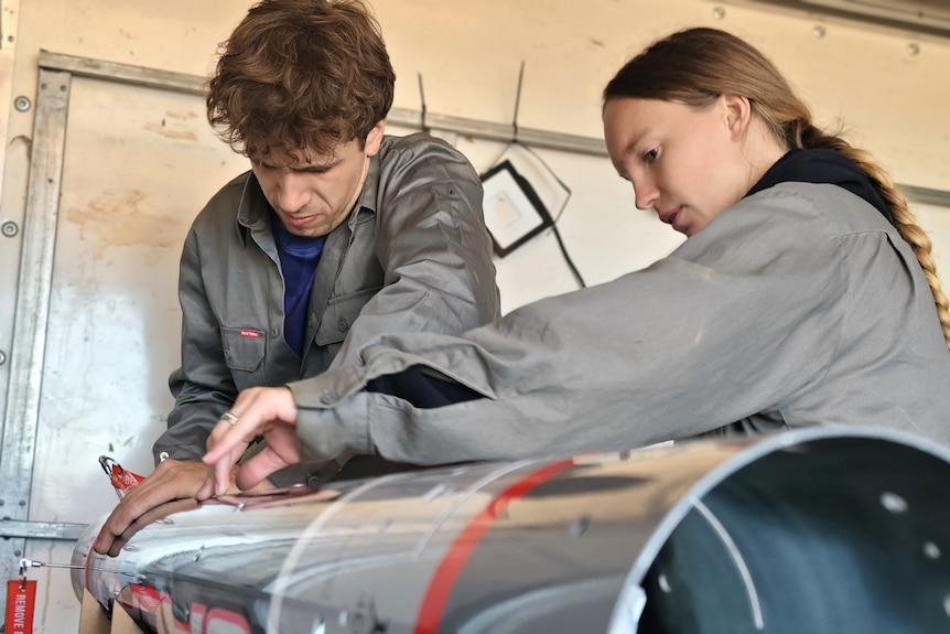 A young man and woman with their hands on a giant grey cylinder, which is an empty rocket. 