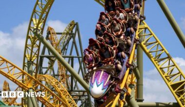 People in a purple rollercoaster race down a yellow track. There are loops and twists on the track behind them.