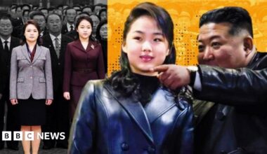 With her long hair tied back, in black trousers and a white padded jacket, Kim Ju Ae walks hand-in-hand with Kim Jong Un, in a brown jacket and black trousers. They are looking towards each other. Towering above them, with several men standing next to it, and hills in the background, an intercontinental ballistic missile (ICBM) points upwards, on a caterpillar-track transporter.