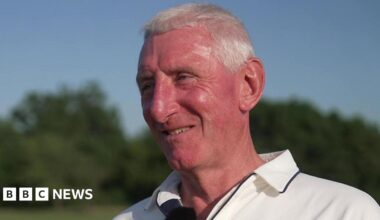 Nic Downes has short white hair and is smiling whilst looking off-camera. He is wearing cricket whites.