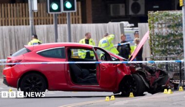 A red car that has crashed has police tape around it. Police officers in the background are wearing neon yellow hi-viz jackets