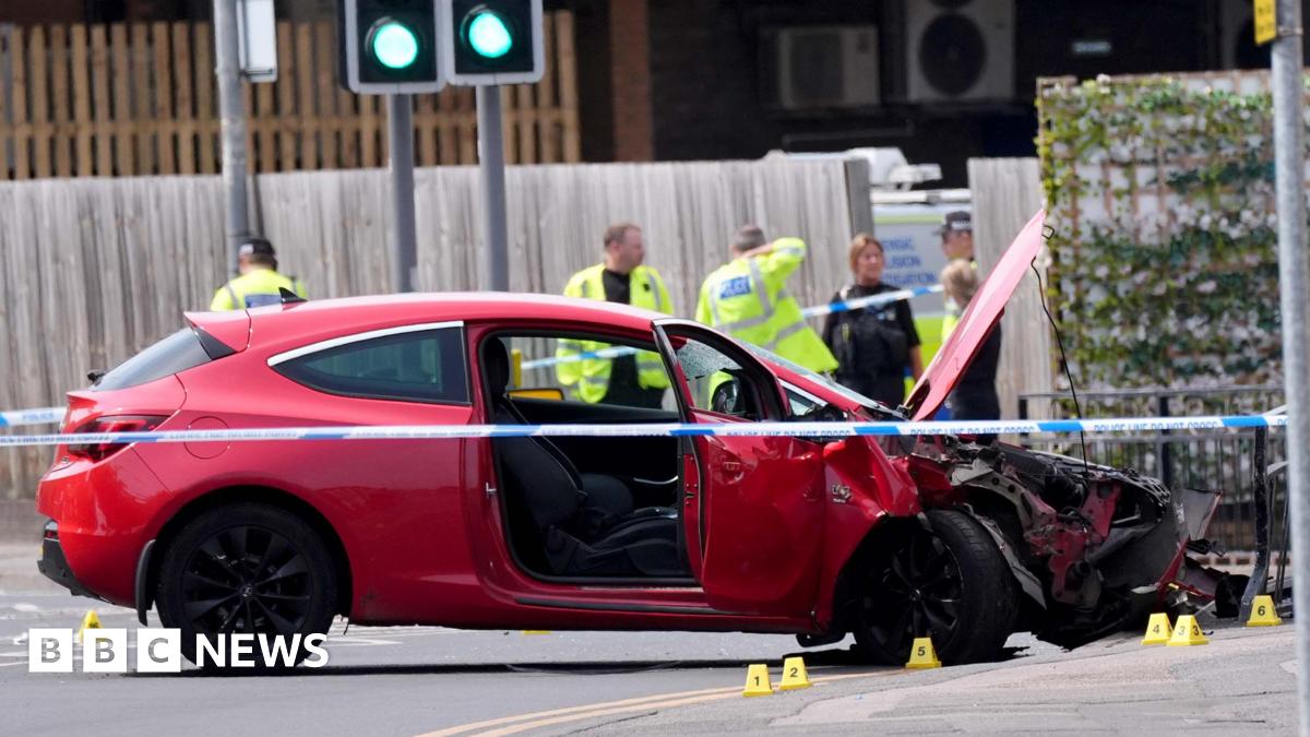 A red car that has crashed has police tape around it. Police officers in the background are wearing neon yellow hi-viz jackets