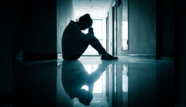 A silhouetted figure sits in a dark hallway with their reflection visible in a shiny floor below them. They are facing to the right and the background has a black and blue colour. Their posture suggests sadness or stress.