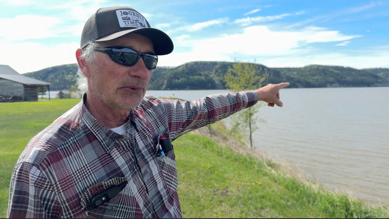a man in a plaid shirt and cap stands on a grassy hill and points toward a big lake reservoir