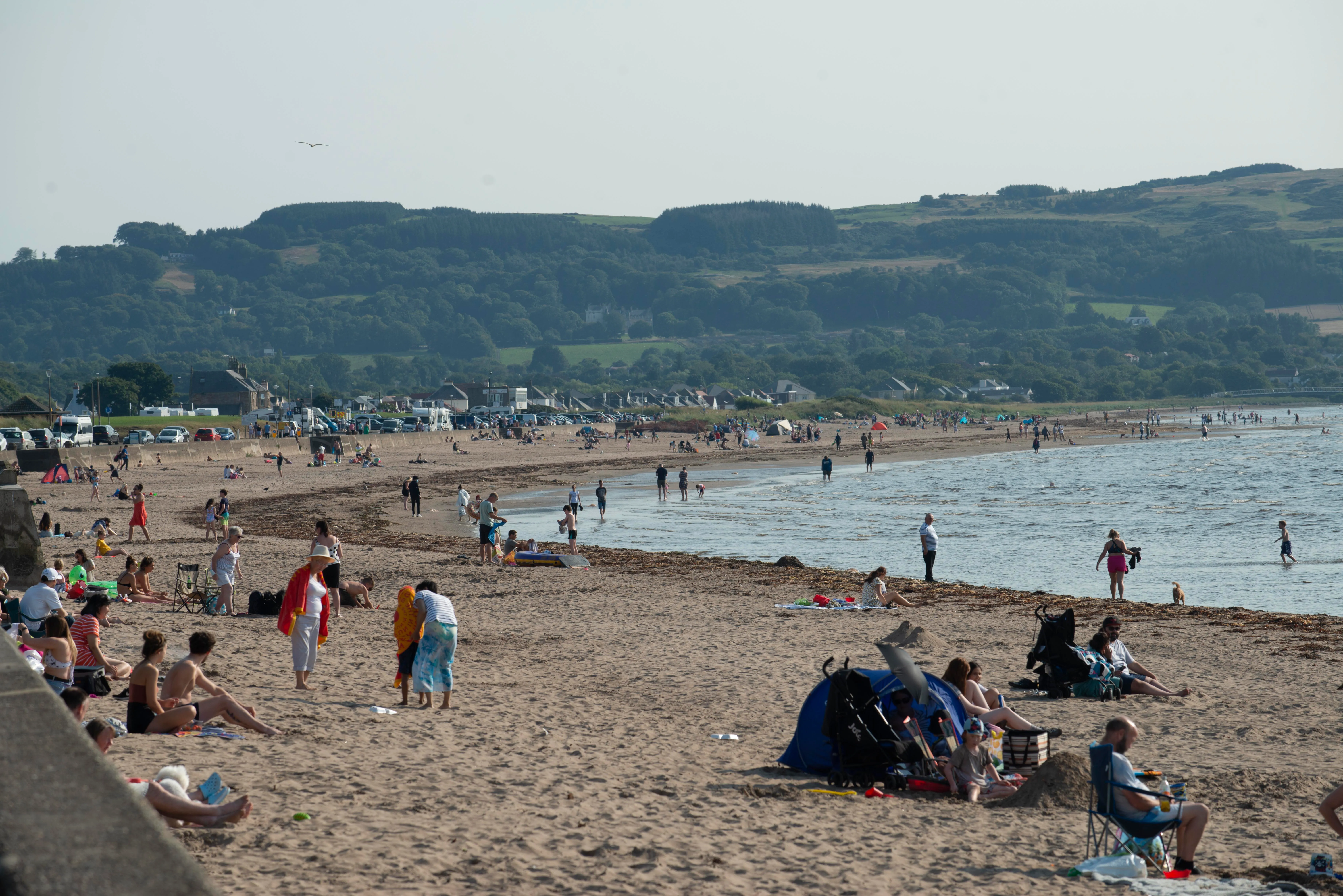People enjoying a day at the beach with a hilly, forested landscape in the background.