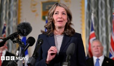 An image of Alberta Premier Danielle Smith standing and speaking at a podium. She is wearing a dark navy blue blazer with a tan top underneath and a silver necklace. She has shoulder-length brown, wavy hair.