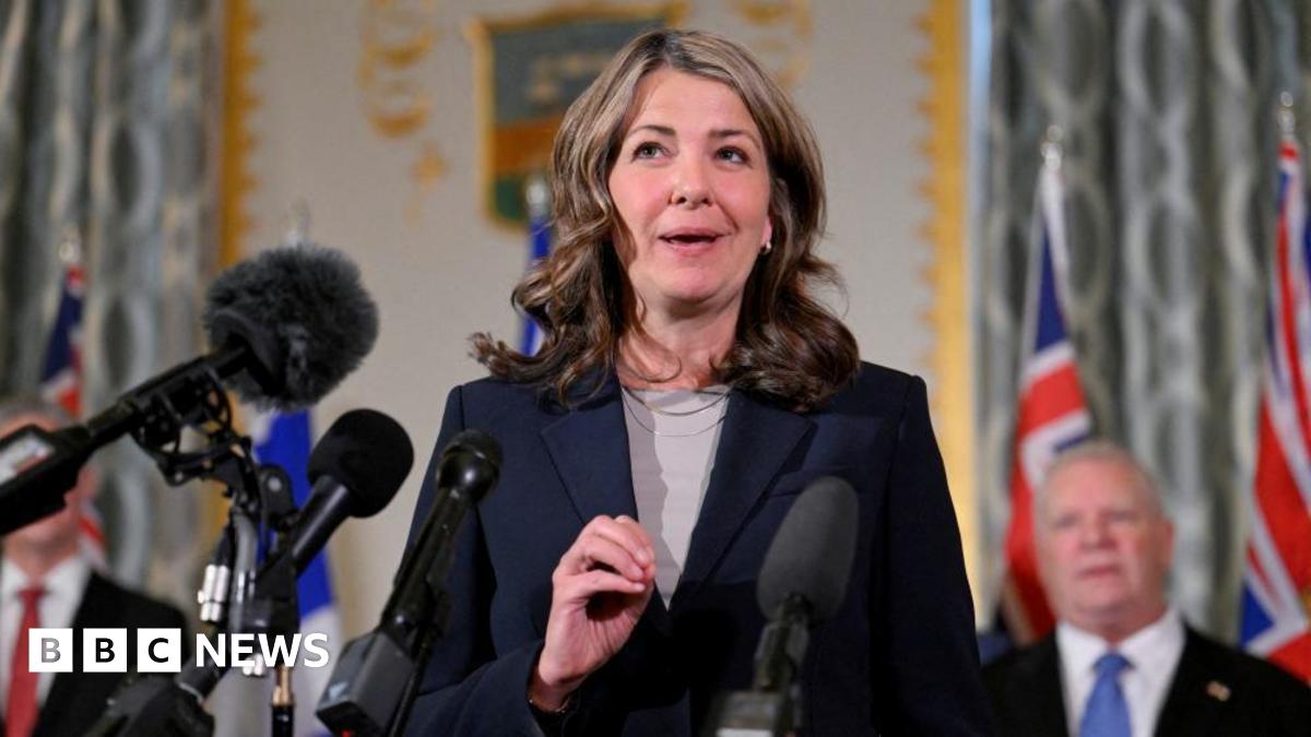 An image of Alberta Premier Danielle Smith standing and speaking at a podium. She is wearing a dark navy blue blazer with a tan top underneath and a silver necklace. She has shoulder-length brown, wavy hair.