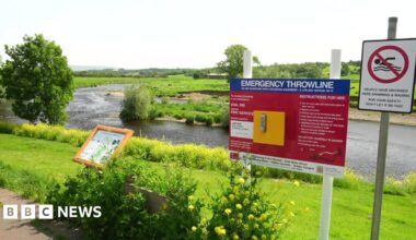 View from the banks of the River Ribble in Ribchester with signs detailing instructions in an emergency and a no swimming sign with a warning saying people have drowned in the river.