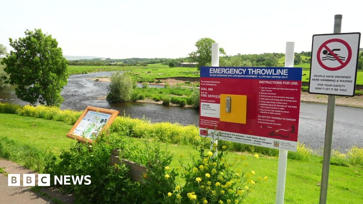 View from the banks of the River Ribble in Ribchester with signs detailing instructions in an emergency and a no swimming sign with a warning saying people have drowned in the river.