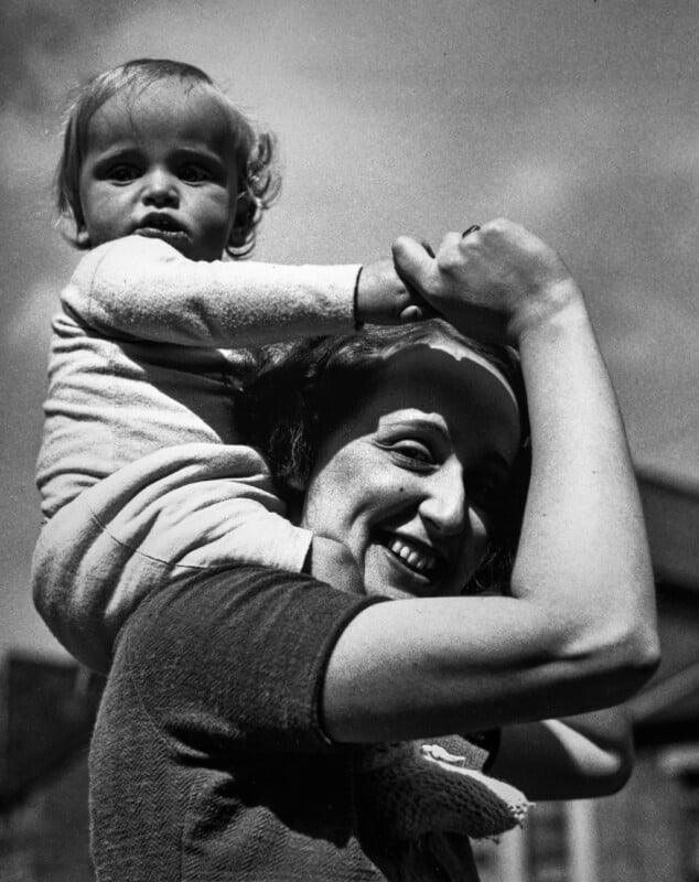A smiling woman carries a young child on her shoulders outdoors. The woman looks up and holds the child's hands while the child gazes ahead. The image is black and white with a clear sky in the background.