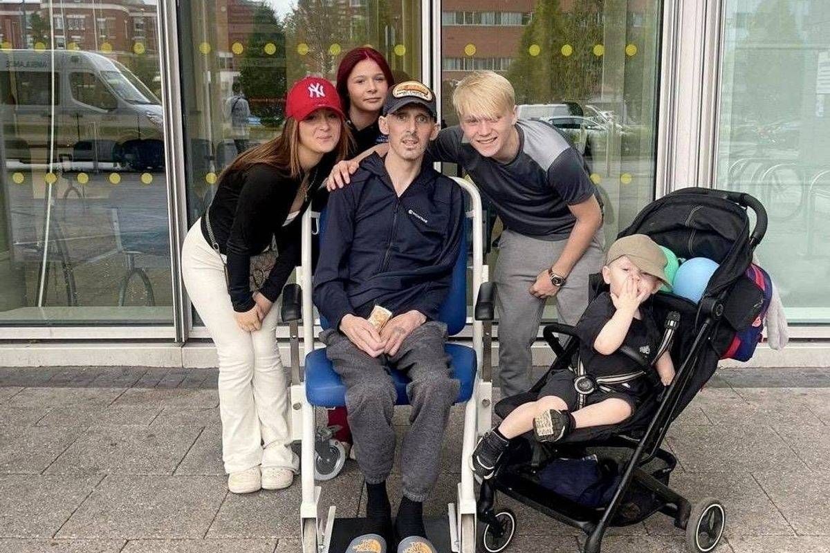 A group of individuals stands together on a paved surface outside a building. A man in the center, seated in a wheelchair, is flanked by a woman in white pants, a man with a hat, and another person. To the right, a child is seated in a stroller. The setting includes a glass wall of the building in the background.