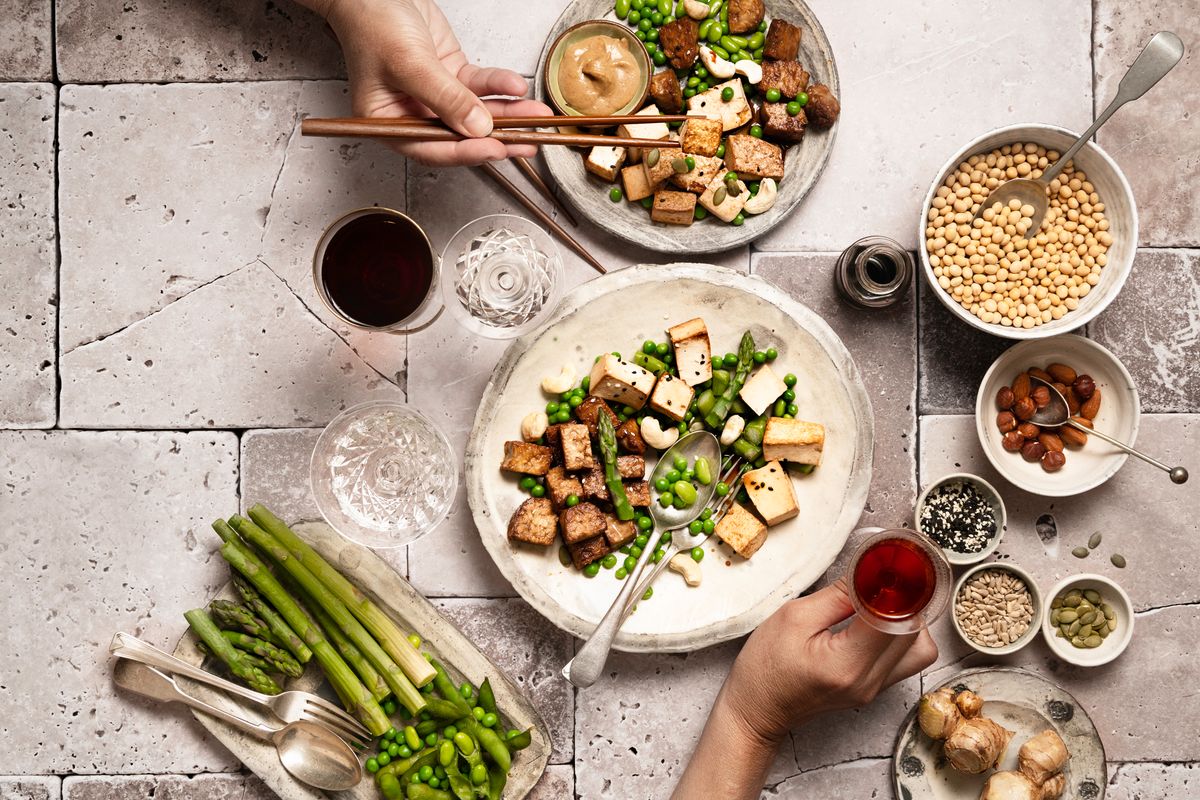 directly above picture of take away vegan and vegetarian foods in plates and bowls with human hands holding glasses, and different bowls containing plant based protein sources, miso sauce, seeds, asparagus, edamame, peas