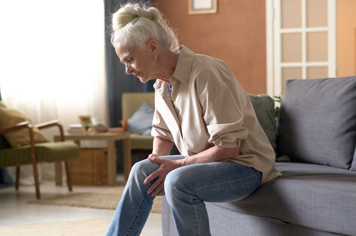 Senior woman sitting on sofa and touching her sore knee, she suffering from serious illness