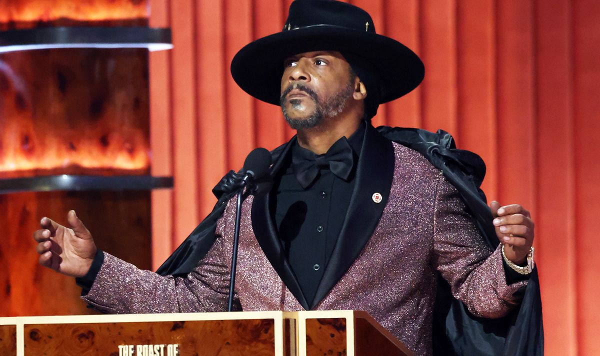 Man in wide-brimmed hat, glittery purple suit jacket and a black cape on stage