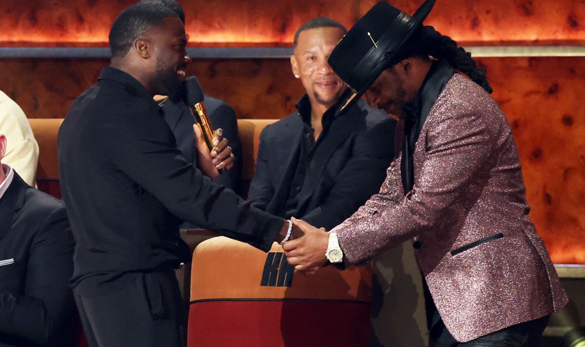 Man in black and man in glittery jacket shake hands and smile on stage