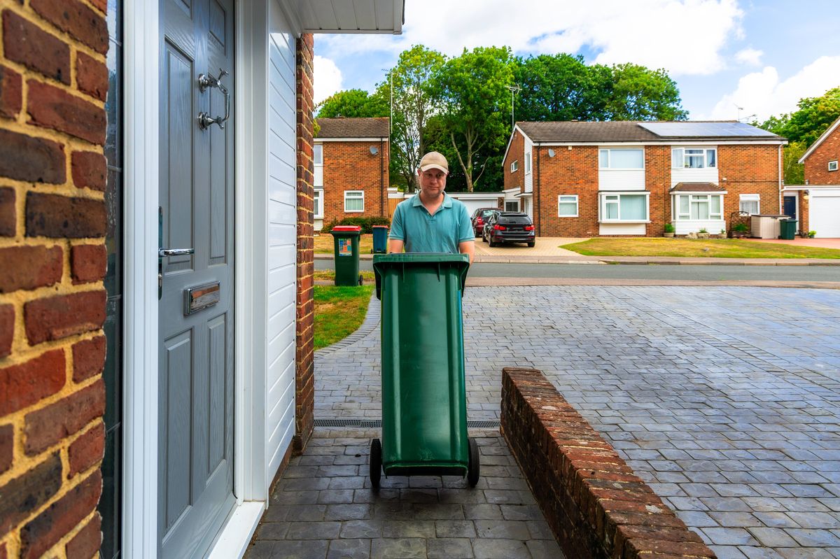 Color image depicting a mid adult male in his 30s taking out his household rubbish bin onto a residential street, awaiting collection. It is summer and the man is dressed casually in beige shorts and red t shirt.