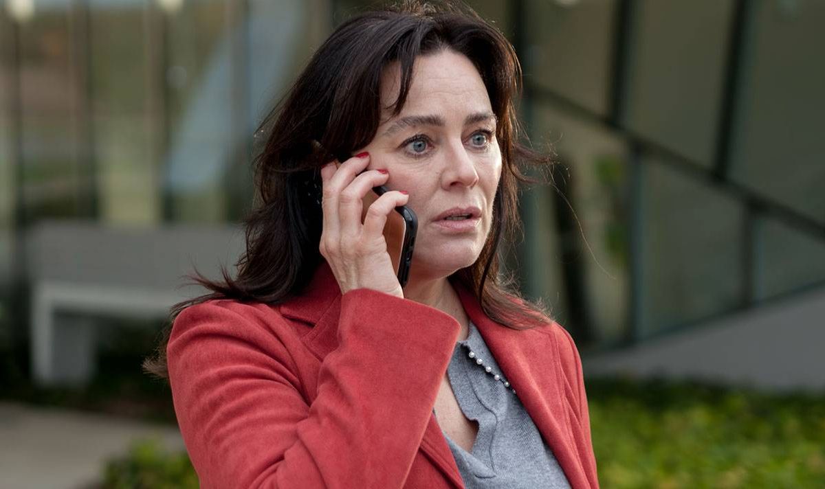 An individual dressed in a red coat and a gray top is engaged in a phone conversation outside a modern building.