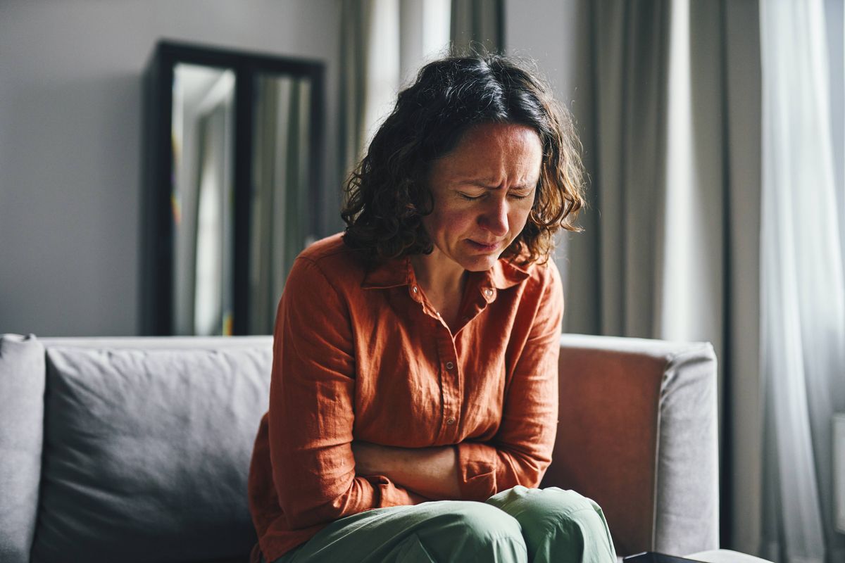 A woman seated indoors, holding her stomach in discomfort and expressing physical distress. The mood captures a realistic depiction of personal pain or health-related issues with natural lighting.
