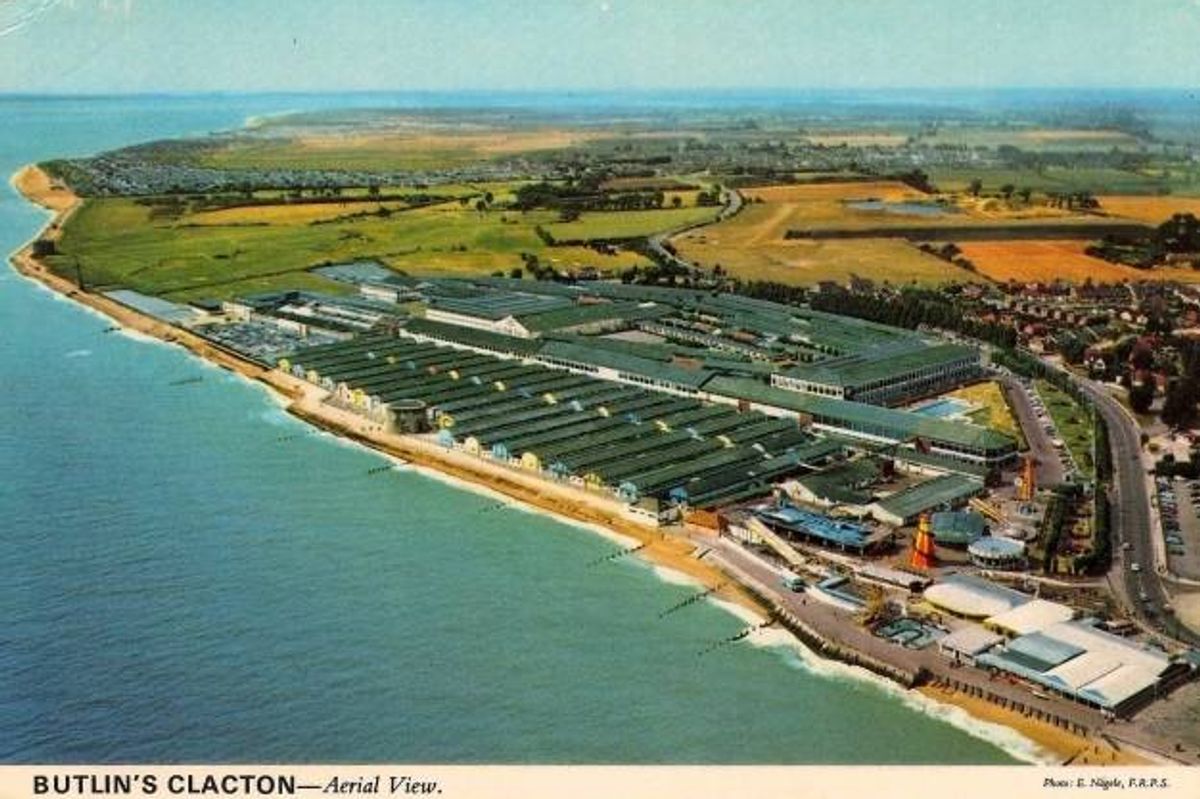 Aerial view of a coastal area featuring a large industrial complex situated along the shoreline. The complex encompasses a substantial area with various structures and facilities visible. To the left, the shoreline curves towards the sea, and to the right, a network of roads and additional buildings is discernible. In the background, a mix of agricultural lands and rural areas can be seen.