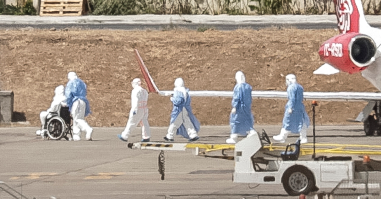 Health personnel in hazmat suits evacuate a passenger in a wheelchair from a cruise ship affected by hantavirus at an airport.