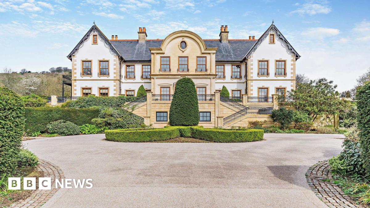 The entrance to a mansion. The property is a beige and white colour with perfectly trimmed bushes in front of the house. A circular driveway can be seen looping around the entrance to the property.