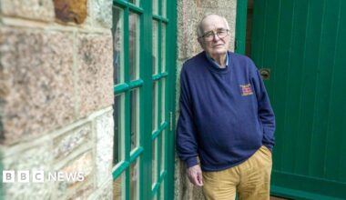 An older man with grey receding hair and glasses leans against a granite wall looking towards the camera.  He wears khaki, flannel trousers and a blue sweatshirt.  He looks sad.