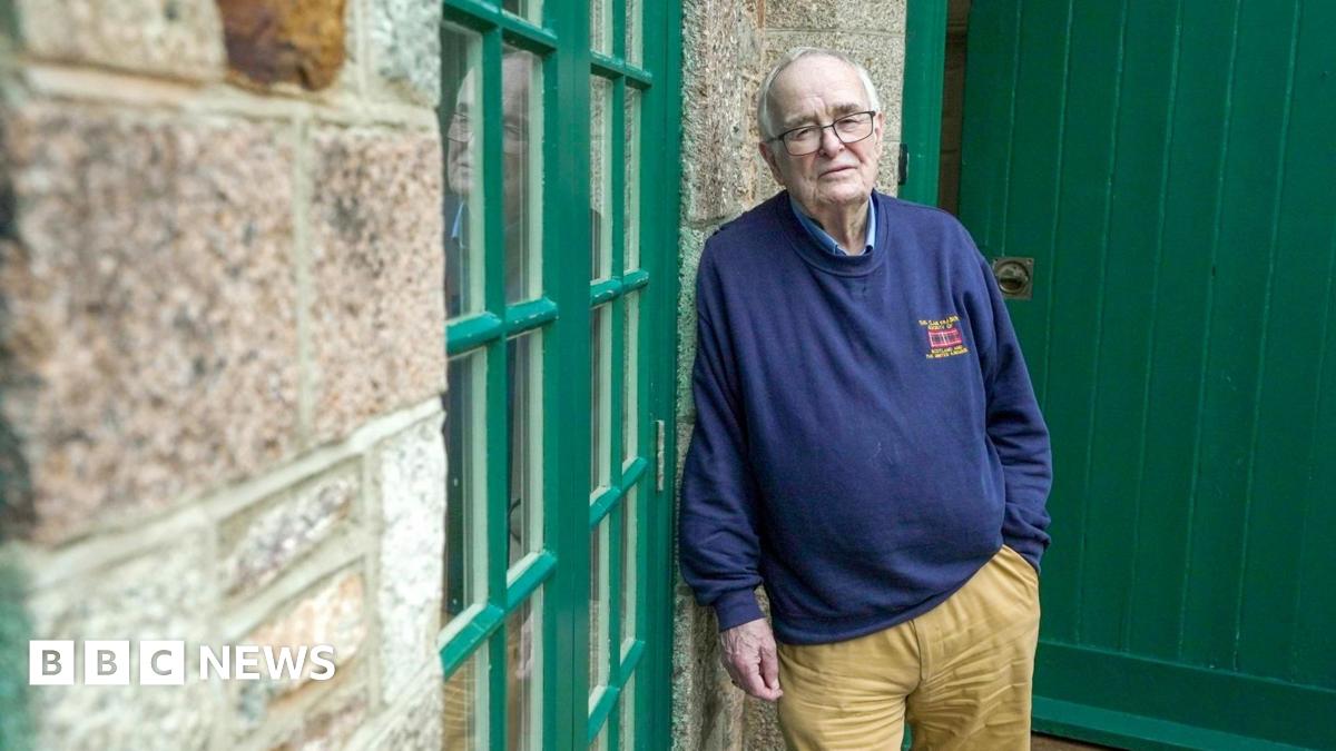 An older man with grey receding hair and glasses leans against a granite wall looking towards the camera.  He wears khaki, flannel trousers and a blue sweatshirt.  He looks sad.