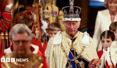 King Charles III is wearing the Imperial State Crown and the Robe of State as he leads the royal procession in the House of Lords. His left hand is held and he has four page boys dressed in red and white to his side.