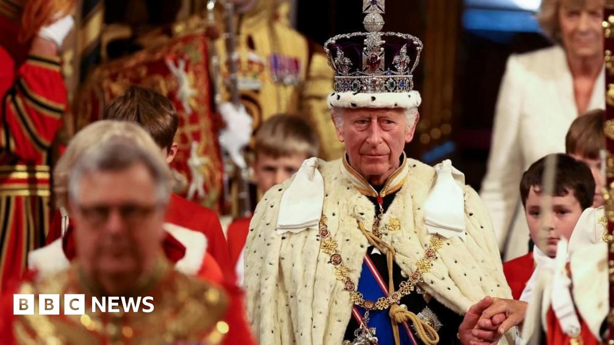 King Charles III is wearing the Imperial State Crown and the Robe of State as he leads the royal procession in the House of Lords. His left hand is held and he has four page boys dressed in red and white to his side.