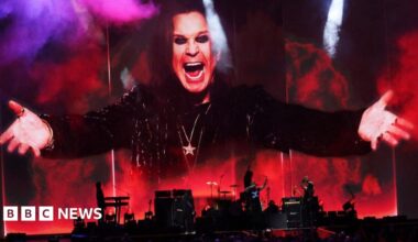 A band can be seen in red and black silouhette at the bottom of the image. On top the image of a man wearing black, chains around his neck, arms outstretched, dark eye make up and with a big smile