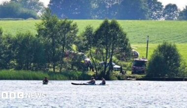 Three men sail canoes on a small lake. Beyone the shoreline opposite to the camera, behind a line of trees a helicopter and emergency service vehicles are visible parked on a grassy slope.