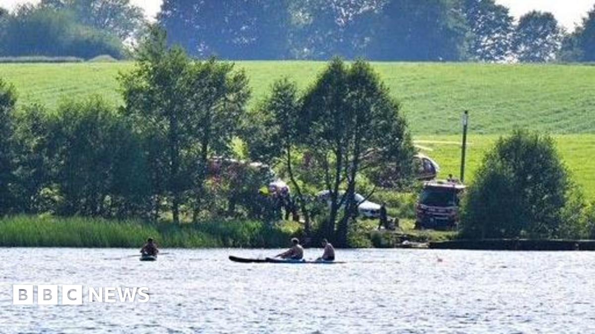 Three men sail canoes on a small lake. Beyone the shoreline opposite to the camera, behind a line of trees a helicopter and emergency service vehicles are visible parked on a grassy slope.