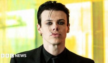 Yungblud looks straight at the camera, wearing a black suit and black tie, against a blurred background.