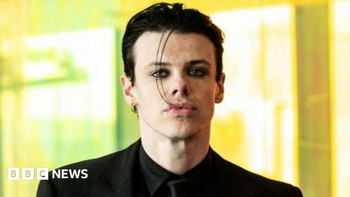 Yungblud looks straight at the camera, wearing a black suit and black tie, against a blurred background.