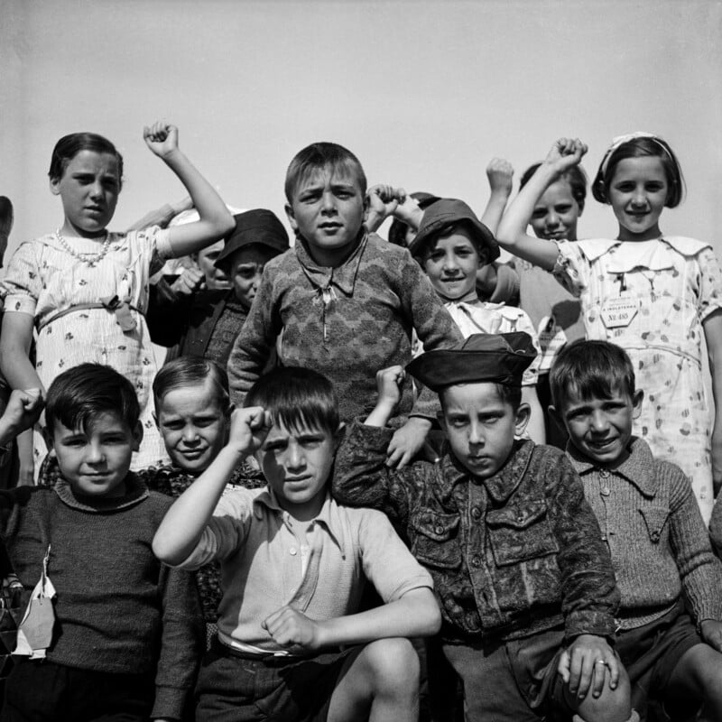 A group of children stand close together outdoors, some raising their fists in the air. They wear a mix of patterned and plain clothes, and most have neutral or serious expressions. The sky behind them is clear.