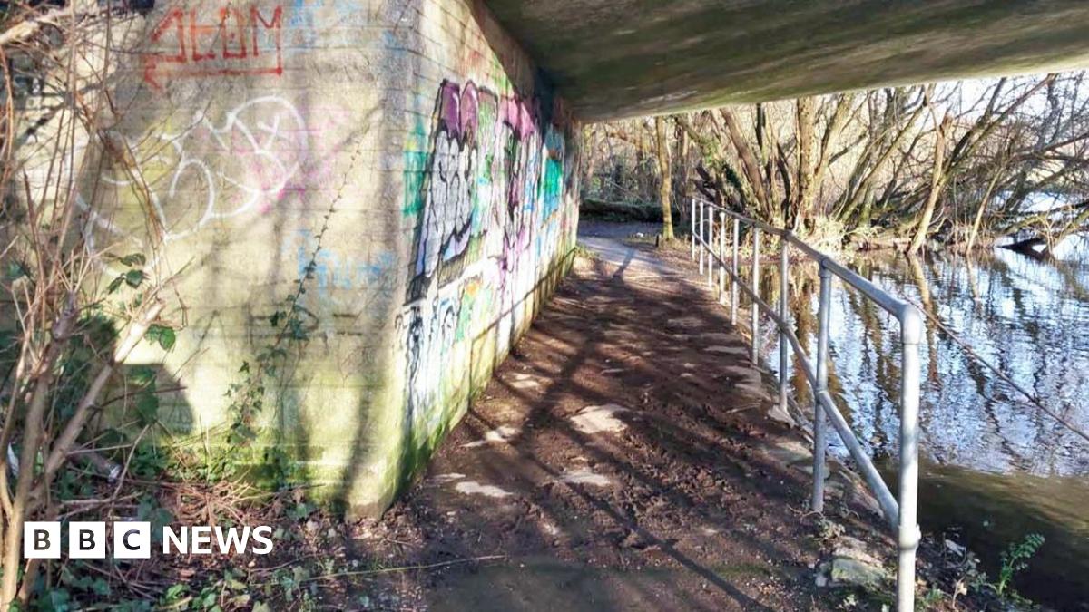 An underpass beneath a bridge next to a river. The wall is covered in graffiti and there is a metal railing along the path.