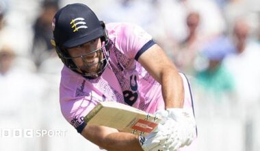 Josh de Caires batting for Middlesex holding his bat out in front of him wearing a pink shirt and helmet