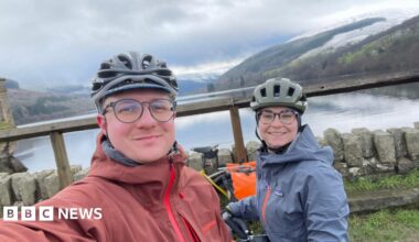 A woman (Margaux Dumas) and a man (Jonas Pollard) both wearing cycling helmets and waterproof jackets. The woman's jacket is blue and the man's is red. In the background is a large body of water and a mountain.