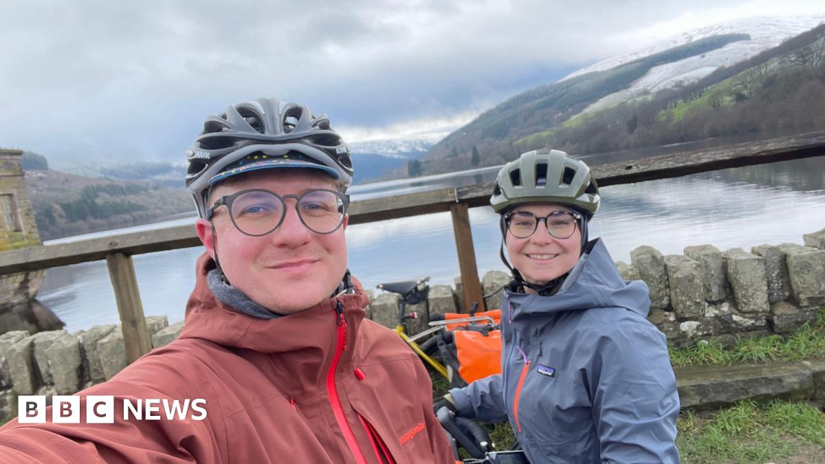 A woman (Margaux Dumas) and a man (Jonas Pollard) both wearing cycling helmets and waterproof jackets. The woman's jacket is blue and the man's is red. In the background is a large body of water and a mountain.
