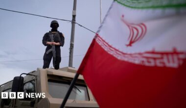 An armed Iranian police officer stands guard on an armored vehicle, monitoring the area during a state-run religious rally in downtown Tehran, Iran, on April 29, 2026. An Iranian flag is seen in the foreground.